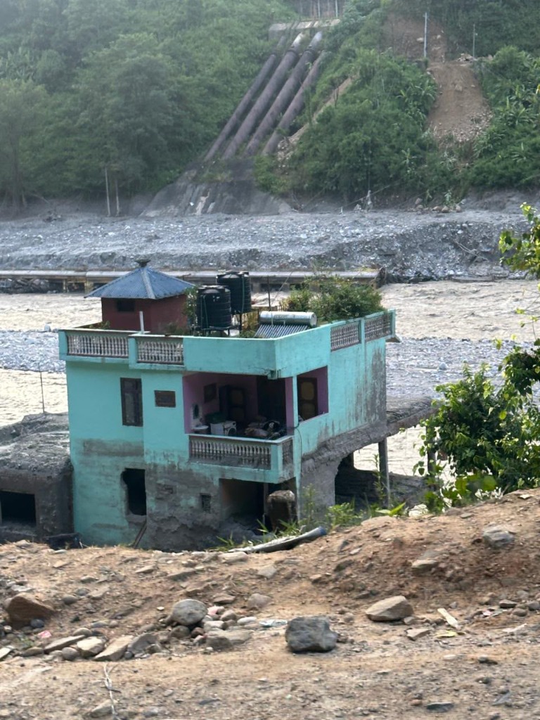 A flood-damaged house standing isolated at the river's edge, its foundation exposed and surrounded by debris and silt